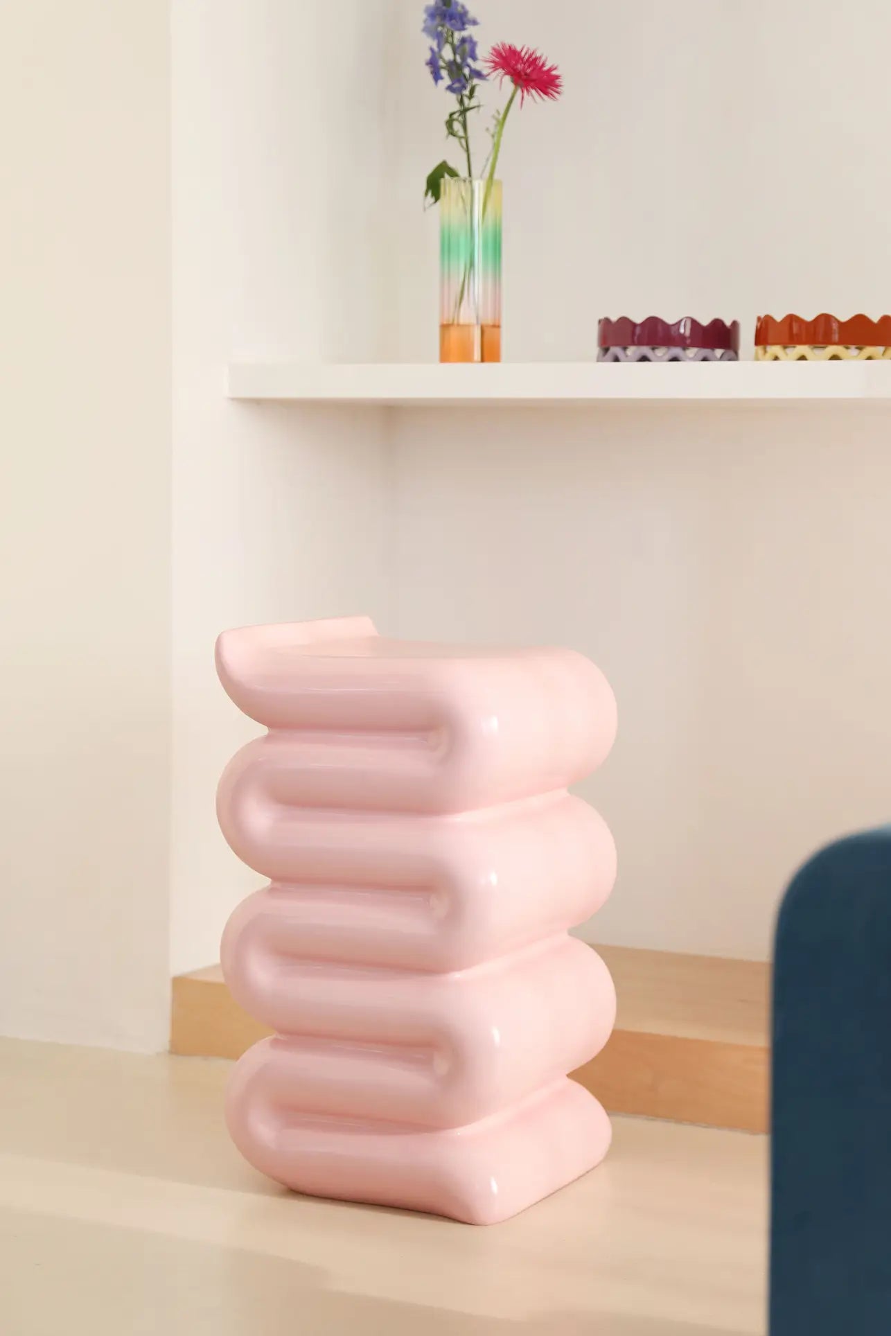 The &klevering Whip Pillar - Pink by &klevering, a pastel sculptural stool with rounded stacked layers, stands on a light floor beside a white shelf that displays a colorful vase with flowers and decorative trays.