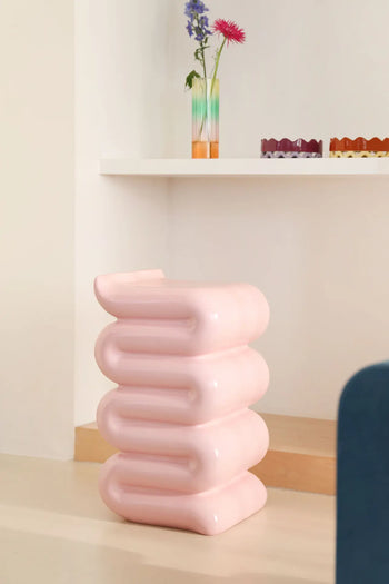 The &klevering Whip Pillar - Pink by &klevering, a pastel sculptural stool with rounded stacked layers, stands on a light floor beside a white shelf that displays a colorful vase with flowers and decorative trays.