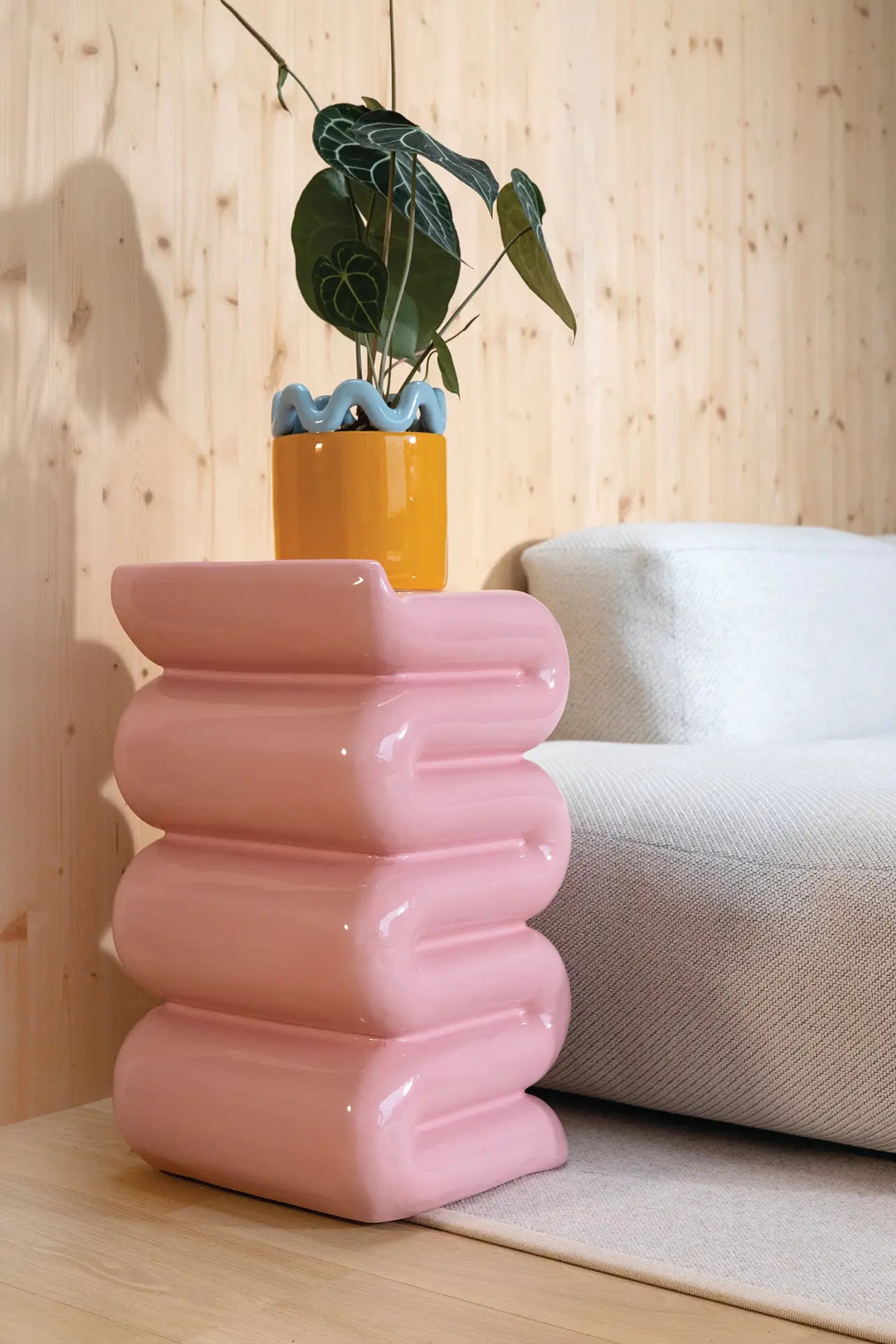 A modern living room featuring the &klevering Whip Pillar - Pink as a sculptural pastel side table, holding a yellow plant pot with wavy blue trim and a green plant, next to a light gray textured sofa and a wooden wall.
