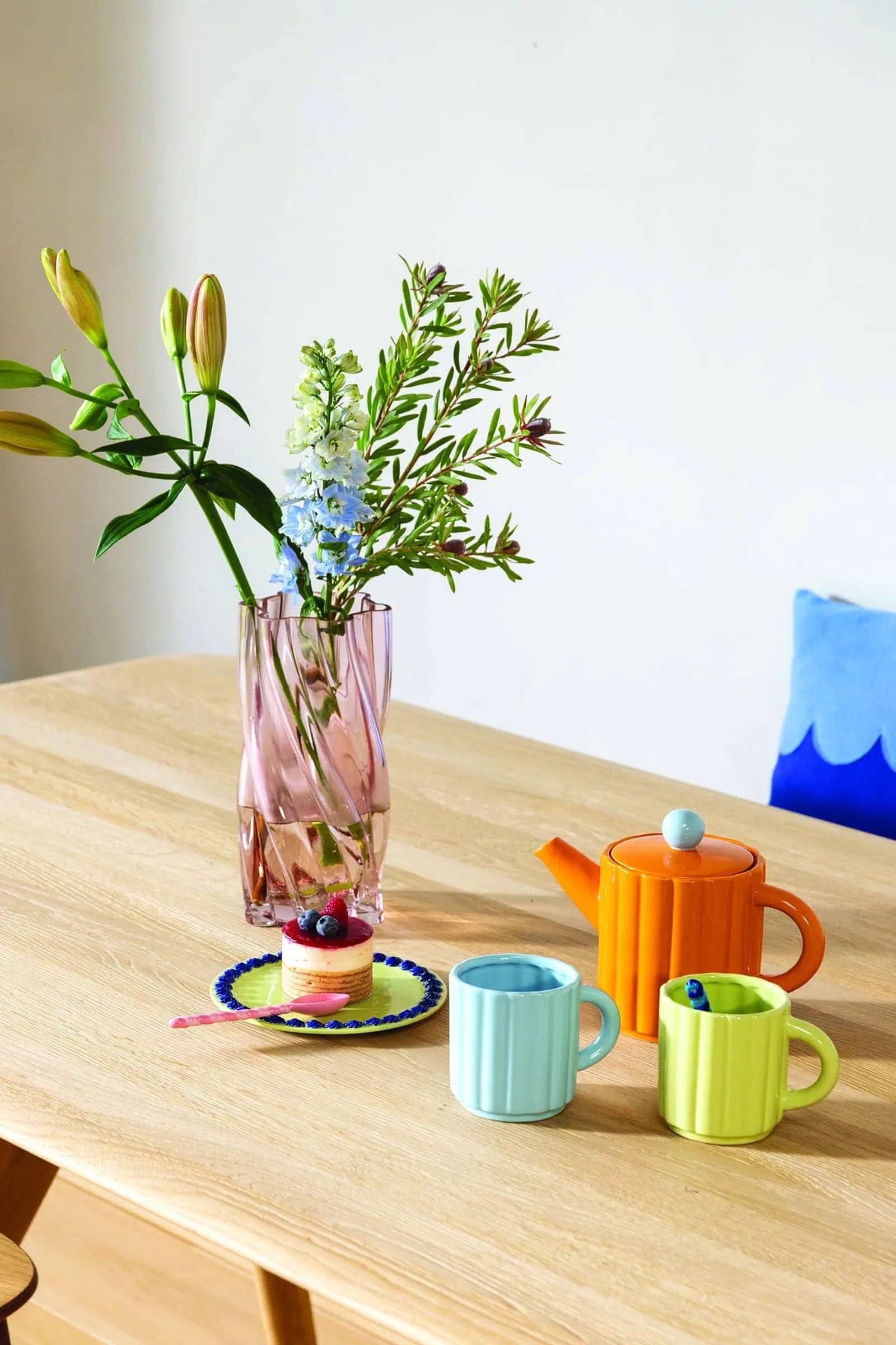 A wooden table in candy colors holds a pink vase with lilies, an orange teapot, blue and green mugs, dessert on the &klevering Whipped Plate (Set of 4), berries, and features a blue pillow in the background.
