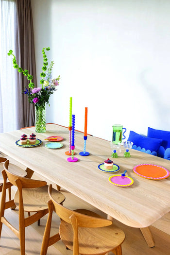 A wooden dining table set with &klevering Whipped Plate, Set of 4 in candy colors, candles, cupcakes with whipped cream, and fresh flowers. Four wooden chairs surround the table; a blue-cushioned bench sits against a white wall.
