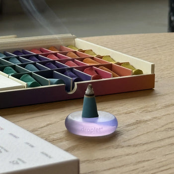 A lit cone from the ripple+ Incense Droplets Multipack sits on a translucent purple holder labeled droplet atop a wooden table, with pastel sticks in a box behind it as fragrant smoke drifts, gently enhancing the mood.