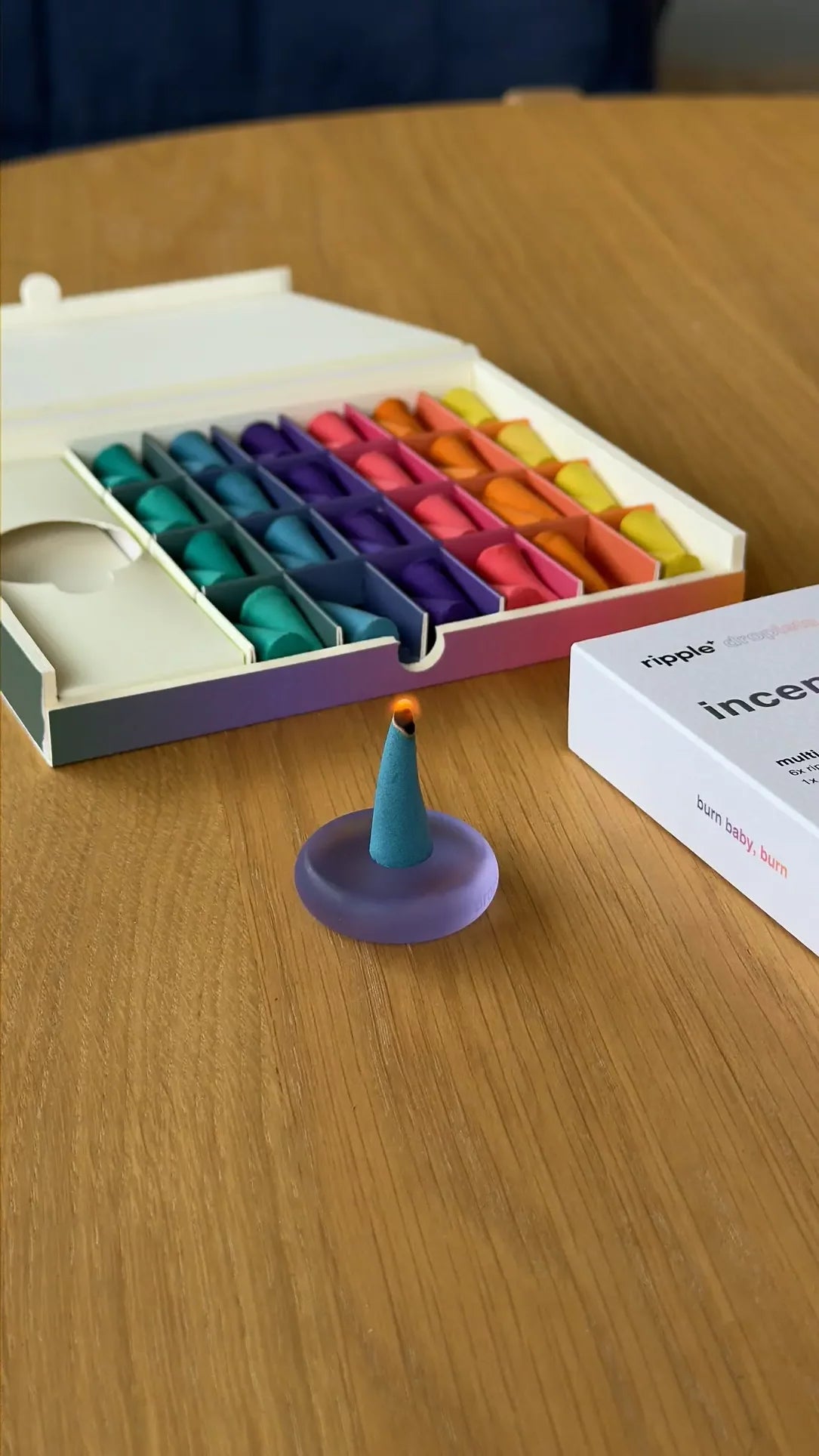 A cone-shaped ripple+ Incense Droplet from the 48-count ripple+ Incense Droplets Multipack sits on a small purple dish, with an open box of colorful droplets and the white packaging box in the background on a wooden table.
