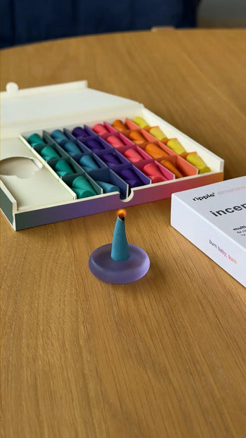A cone-shaped ripple+ Incense Droplet from the 48-count ripple+ Incense Droplets Multipack sits on a small purple dish, with an open box of colorful droplets and the white packaging box in the background on a wooden table.