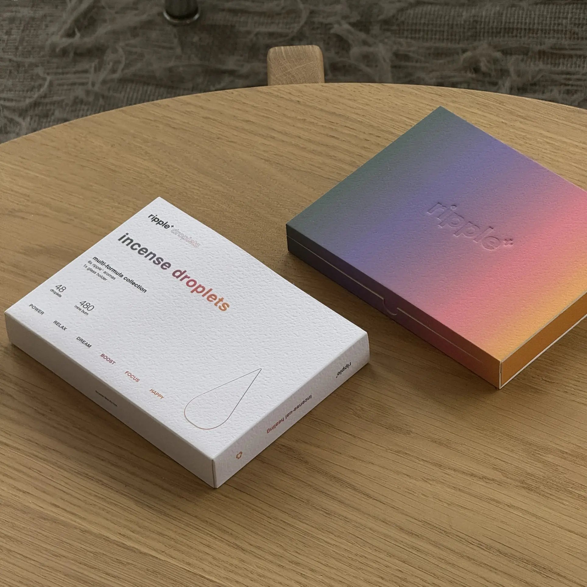 Two square boxes rest on a light wooden table: one, labeled ripple+ Incense Droplets Multipack, 48 Droplets, suggests aromatic scents by ripple+, while the other is iridescent with embossed “ripple+.” A carpet appears in the background.
