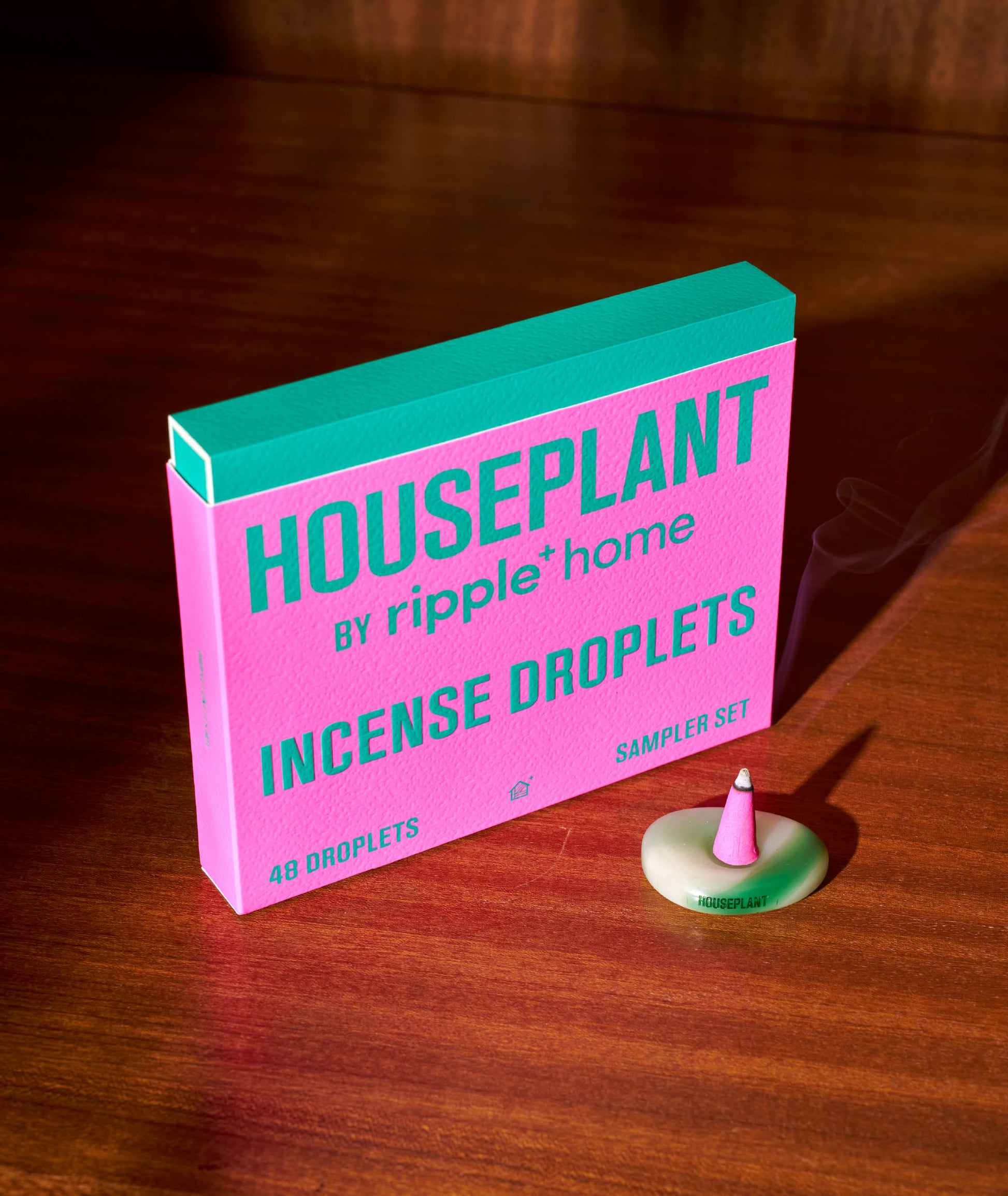 A ripple+ Houseplant Incense Droplets Sampler Set in pink and green rests on a wooden surface beside a hand-rolled incense cone on a white dish, gently releasing aromatic smoke.
