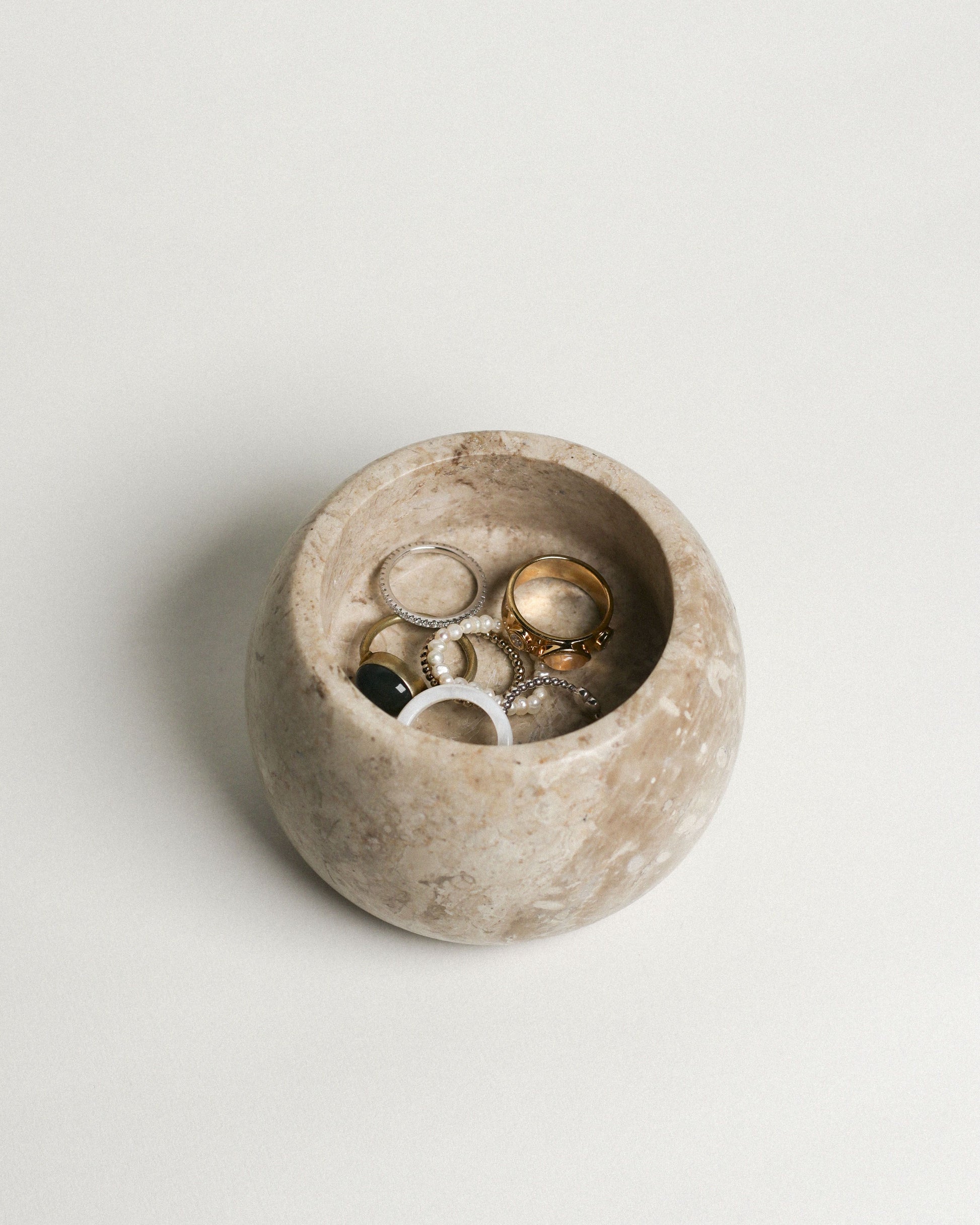 The stillgoods Abel Duo Holder, a round beige stone storage dish by stillgoods, displays assorted rings—gold, silver, and gemstone bands—arranged on a plain light surface.