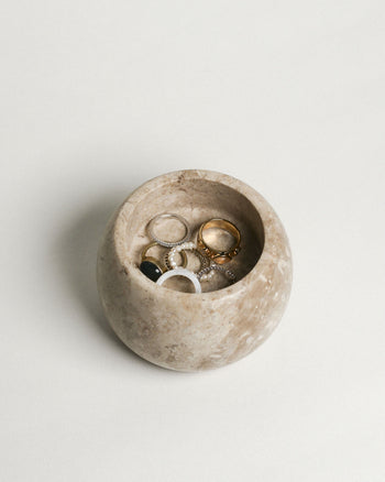 The stillgoods Abel Duo Holder, a round beige stone storage dish by stillgoods, displays assorted rings—gold, silver, and gemstone bands—arranged on a plain light surface.