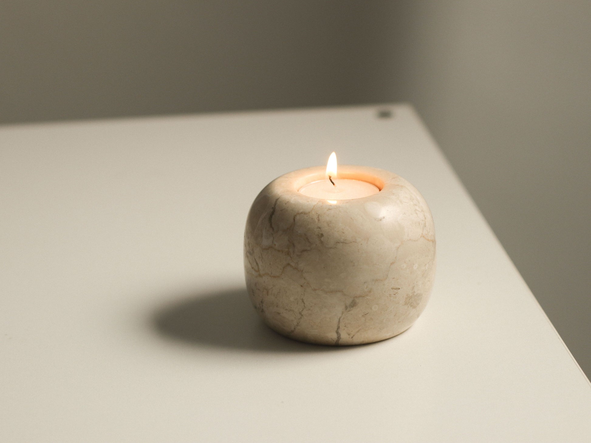 The stillgoods Abel Duo Holder, a small beige marble tealight candle holder with a lit candle, sits on a smooth light surface and casts a soft shadow in a minimalist setting.