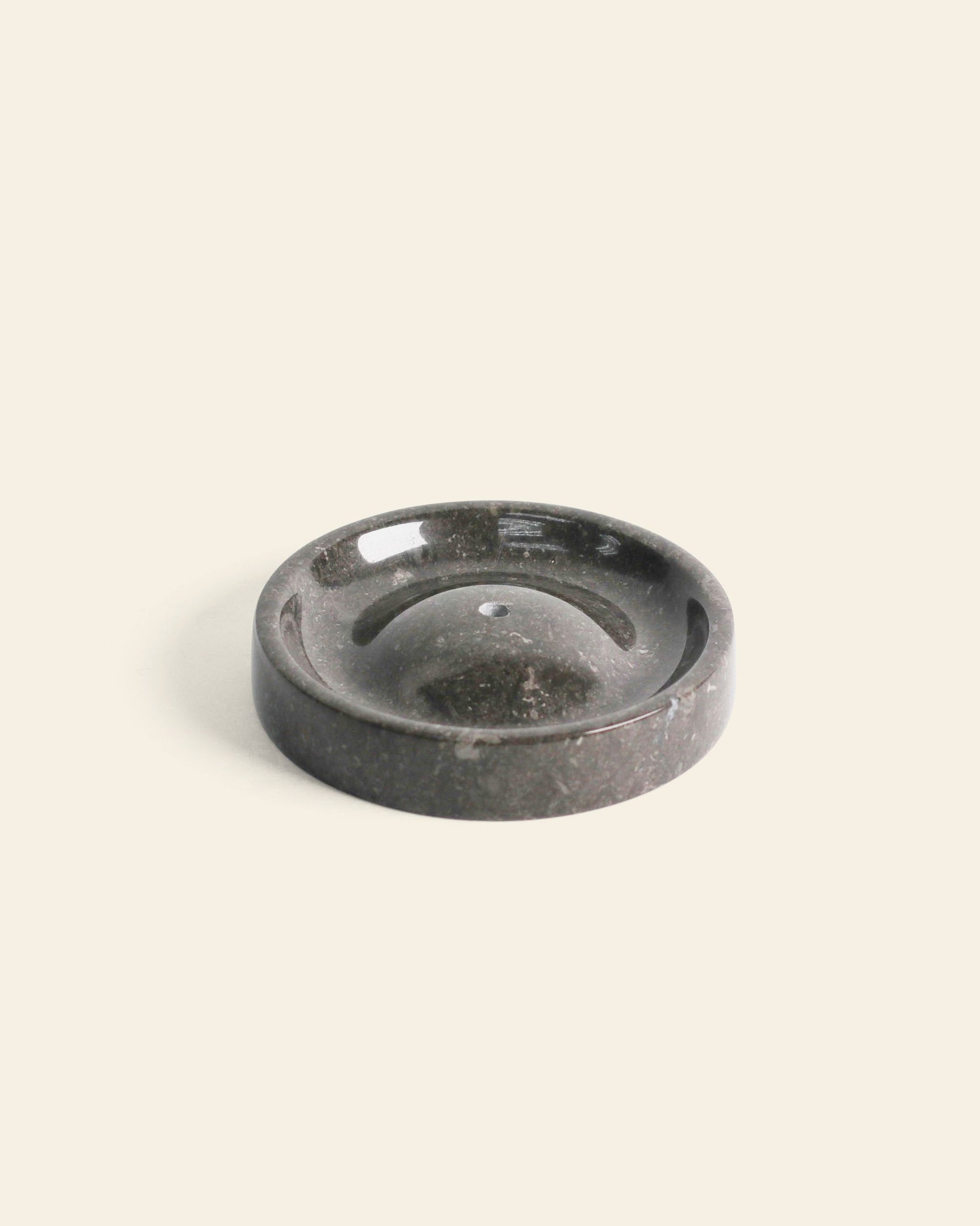 The stillgoods Ebb Incense Holder features a round, dark marble design with a central hole and recessed groove, set against a light background. Its minimalist style works as both an incense holder and an elegant accessory tray.
