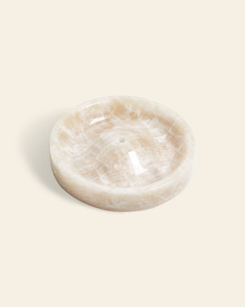 The stillgoods Ebb Incense Holder is a round, shallow tray crafted from light beige marble with subtle veining, a smooth polished finish, and a central hole—ideal for ikebana or incense—shown against an off-white background.