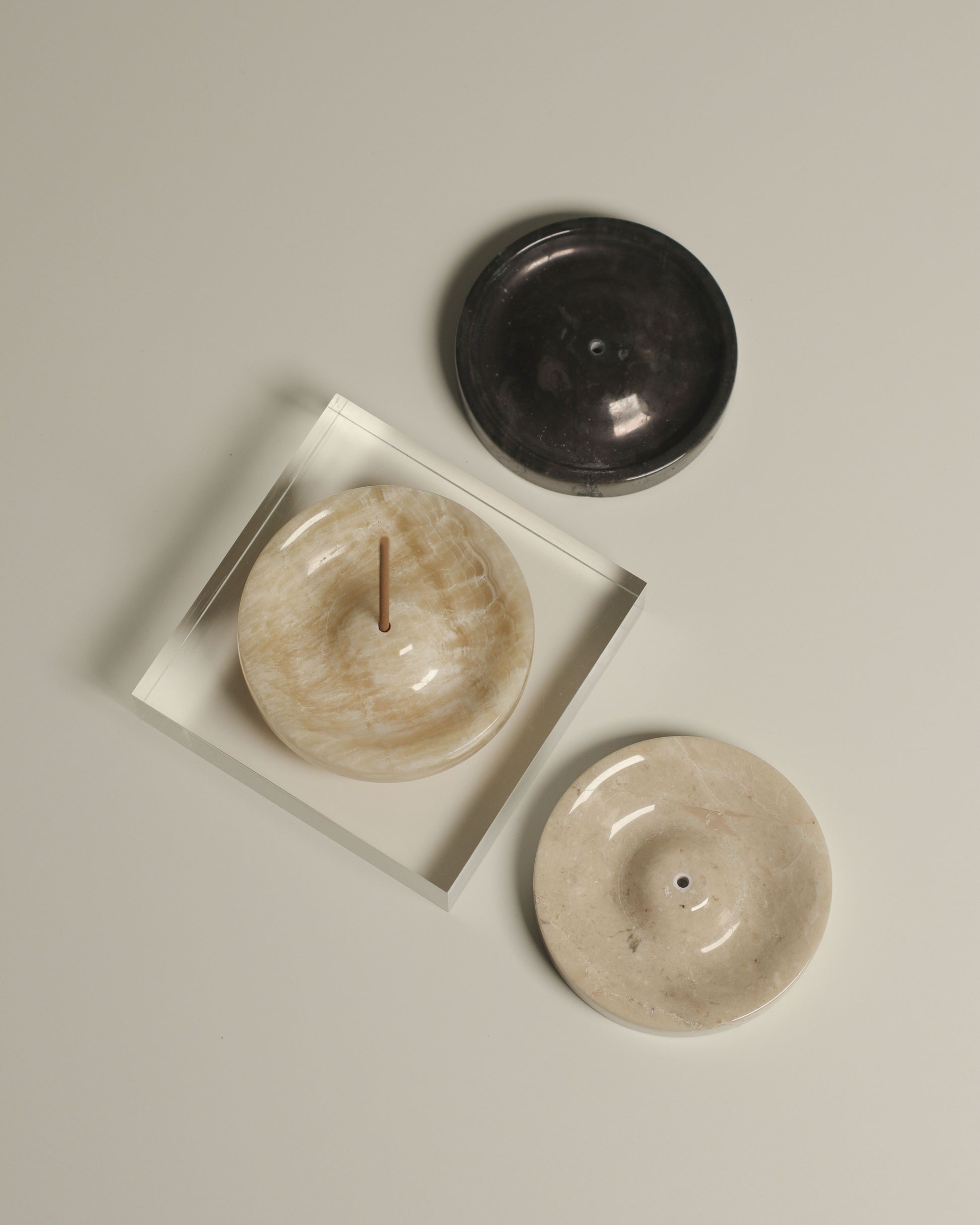 Three stillgoods Ebb Incense Holders in black, beige, and cream are displayed on a light surface. The cream holder doubles as an incense and ikebana holder, shown with burning incense in a clear tray. Brand: stillgoods.