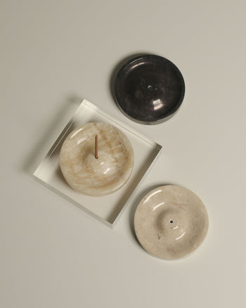 Three stillgoods Ebb Incense Holders in black, beige, and cream are displayed on a light surface. The cream holder doubles as an incense and ikebana holder, shown with burning incense in a clear tray. Brand: stillgoods.