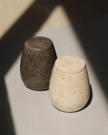 Two stillgoods jars—one Ecru Jar and one dark brown—sit side by side on a light surface, partly in soft shadow. Ideal as versatile organizers or for stylish condiment storage, these rounded stone containers come with matching lids.