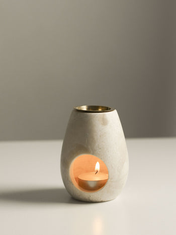 A lit tea light glows inside the stillgoods Eli Oil Burner, diffusing essential oils and filling the room with a calming scent. Its smooth, rounded design and open window cast warmth on a white surface against a neutral backdrop.