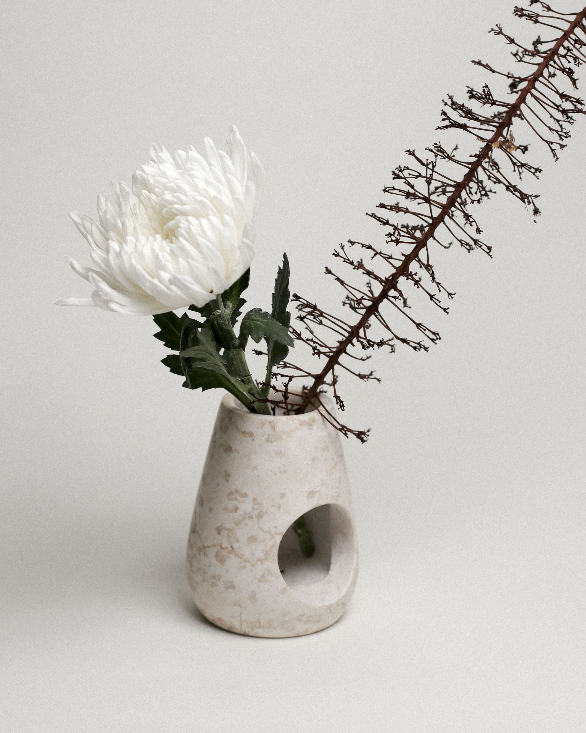 A minimalist beige vase, inspired by the stillgoods Eli Oil Burner, holds a single white chrysanthemum and a dried brown branch against a plain light background.