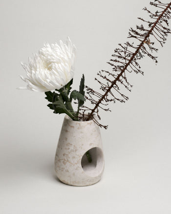 A minimalist beige vase, inspired by the stillgoods Eli Oil Burner, holds a single white chrysanthemum and a dried brown branch against a plain light background.