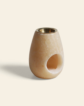The stillgoods Eli Oil Burner is a smooth, pale beige stone piece with a rounded shape, brass top insert, and side circular cutout—ideal for calming scents—shown against a plain light background.