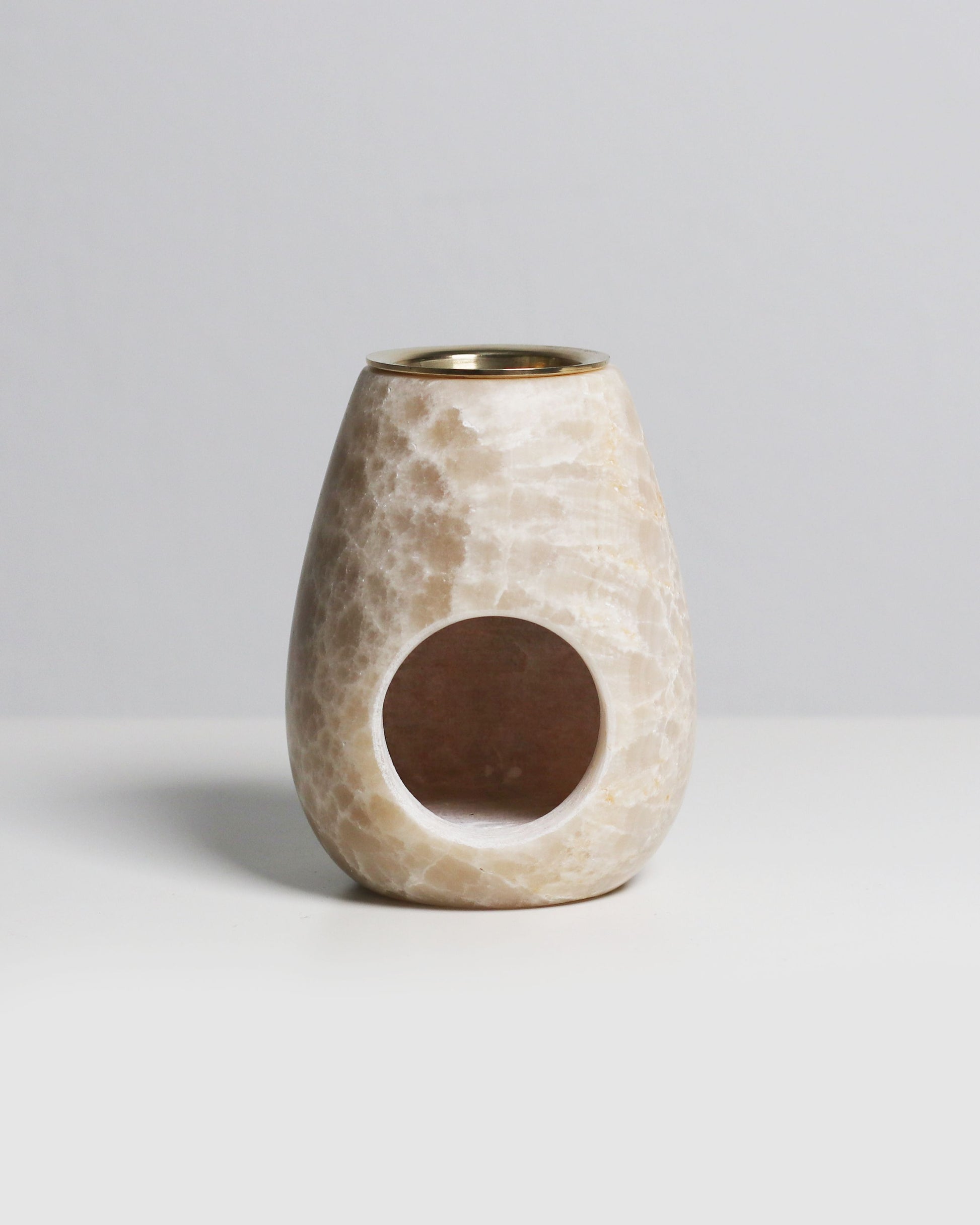 The stillgoods Eli Oil Burner is a beige, marble-patterned, oval essential oil diffuser with a circular front opening, ideal for adding calming scents to any space. Displayed on a white surface with a light gray background.