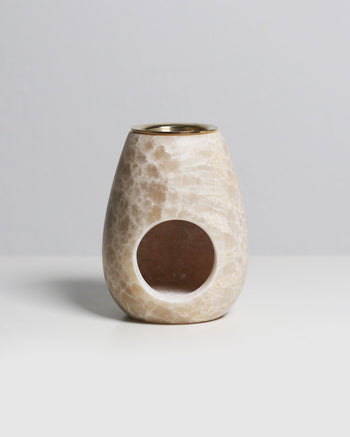 The stillgoods Eli Oil Burner is a beige, marble-patterned, oval essential oil diffuser with a circular front opening, ideal for adding calming scents to any space. Displayed on a white surface with a light gray background.