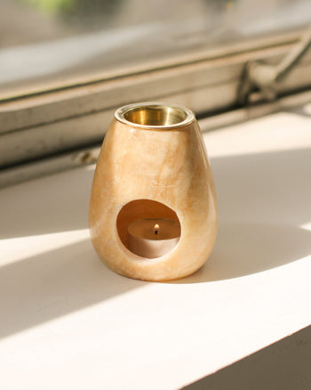 A small, beige stillgoods Eli Oil Burner with a rounded shape sits on a white windowsill, holding a lit tealight candle. Sunlight streams in, casting gentle shadows and suggesting the soothing aroma of this essential oil diffuser.