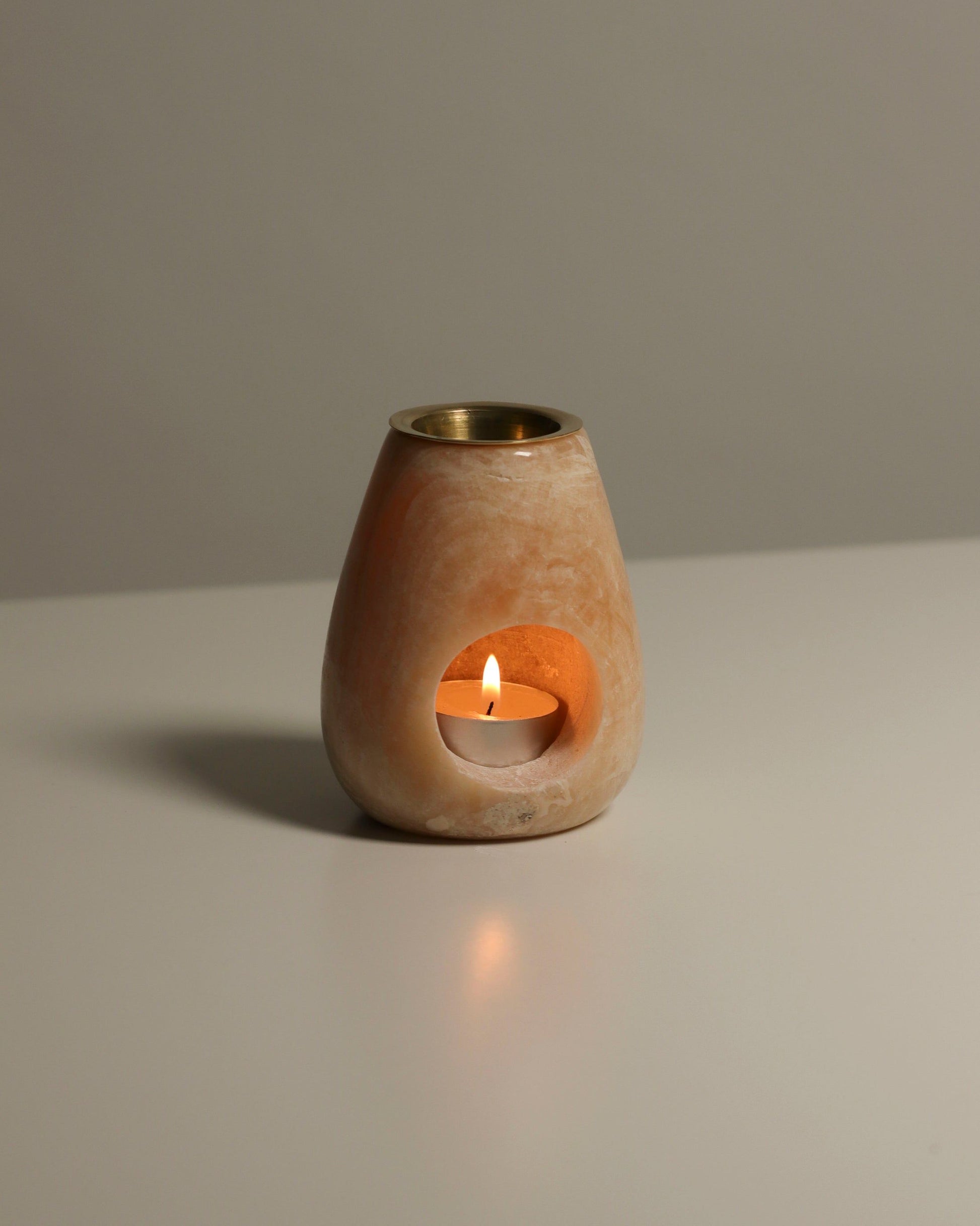 A lit tealight sits inside the stillgoods Eli Oil Burner, a beige stone holder with a round opening, casting a warm glow on a white surface and ready to fill the room with calming fragrance.
