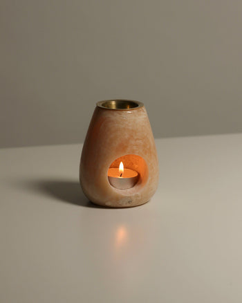 A lit tealight sits inside the stillgoods Eli Oil Burner, a beige stone holder with a round opening, casting a warm glow on a white surface and ready to fill the room with calming fragrance.