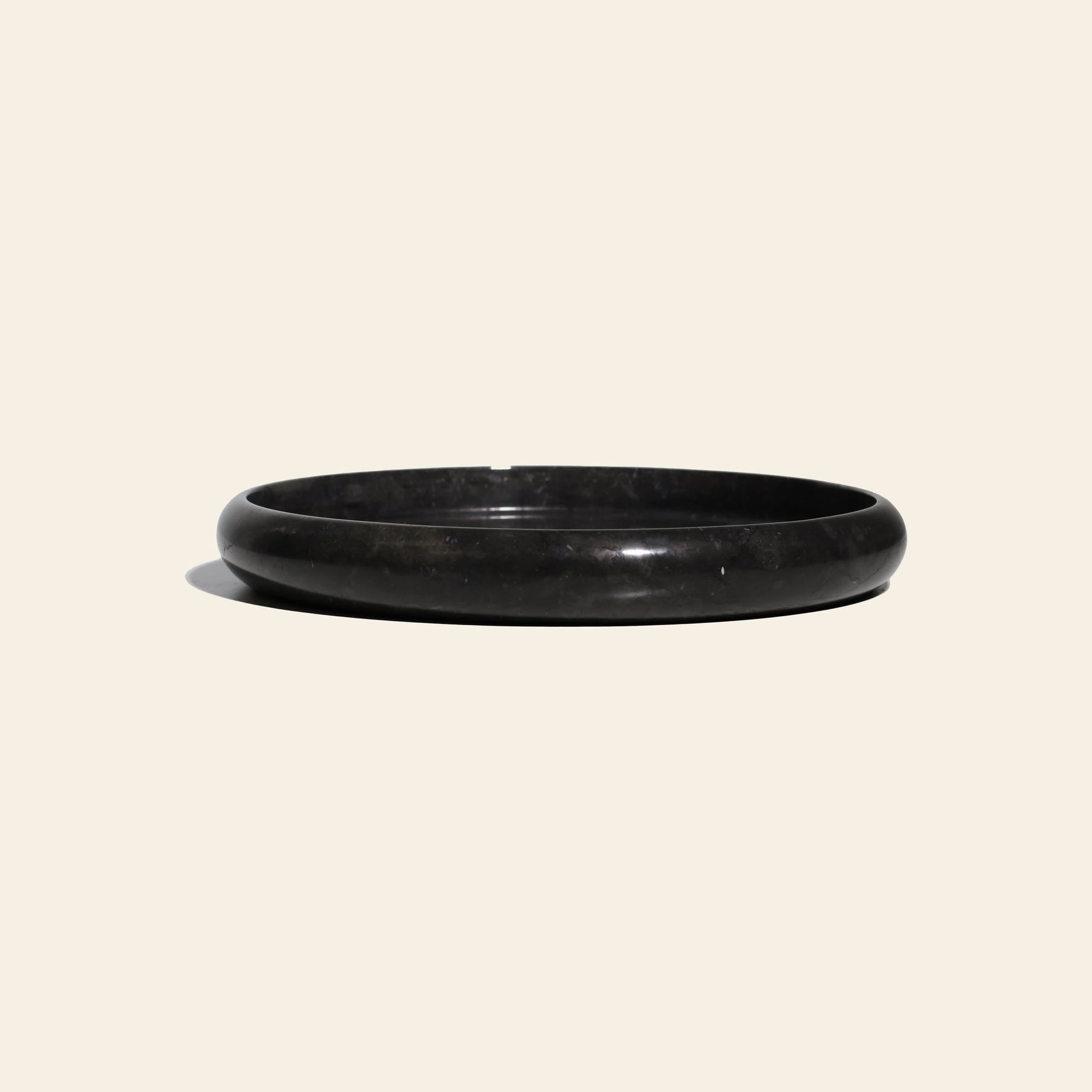 The stillgoods Harmony Tray by stillgoods is a round, shallow black stone tray with a smooth surface and slightly raised rim, ideal as a marble centerpiece for decor, shown on a light beige background with a soft shadow on the left.