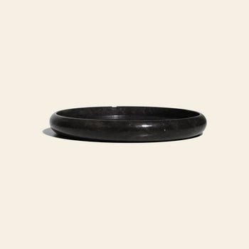The stillgoods Harmony Tray by stillgoods is a round, shallow black stone tray with a smooth surface and slightly raised rim, ideal as a marble centerpiece for decor, shown on a light beige background with a soft shadow on the left.