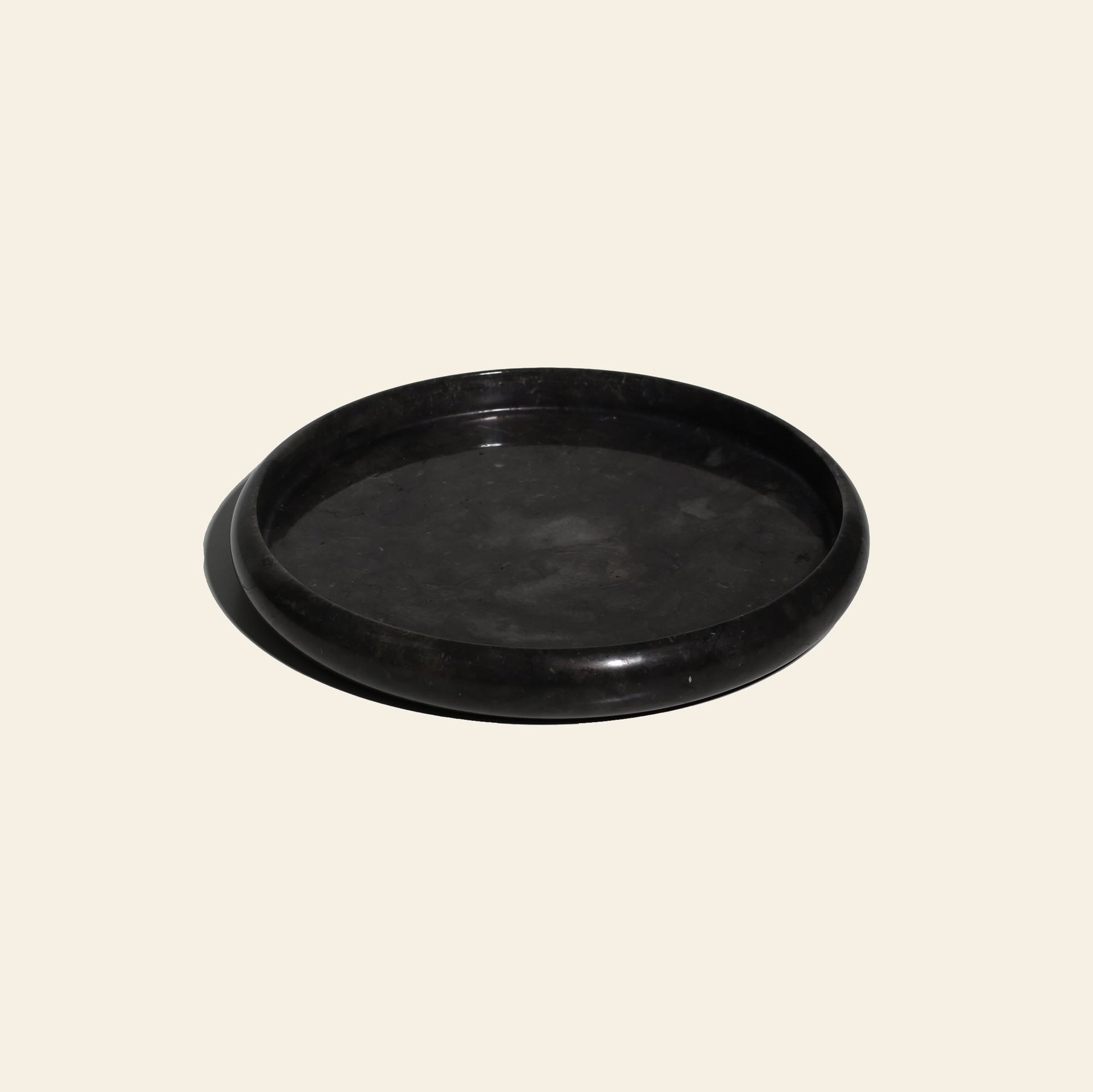 The stillgoods Harmony Tray is a round, flat black stone tray with gently raised edges. Perfect as a chic marble centerpiece to elevate any decor, it sits on a light beige background with a subtle shadow to the left.