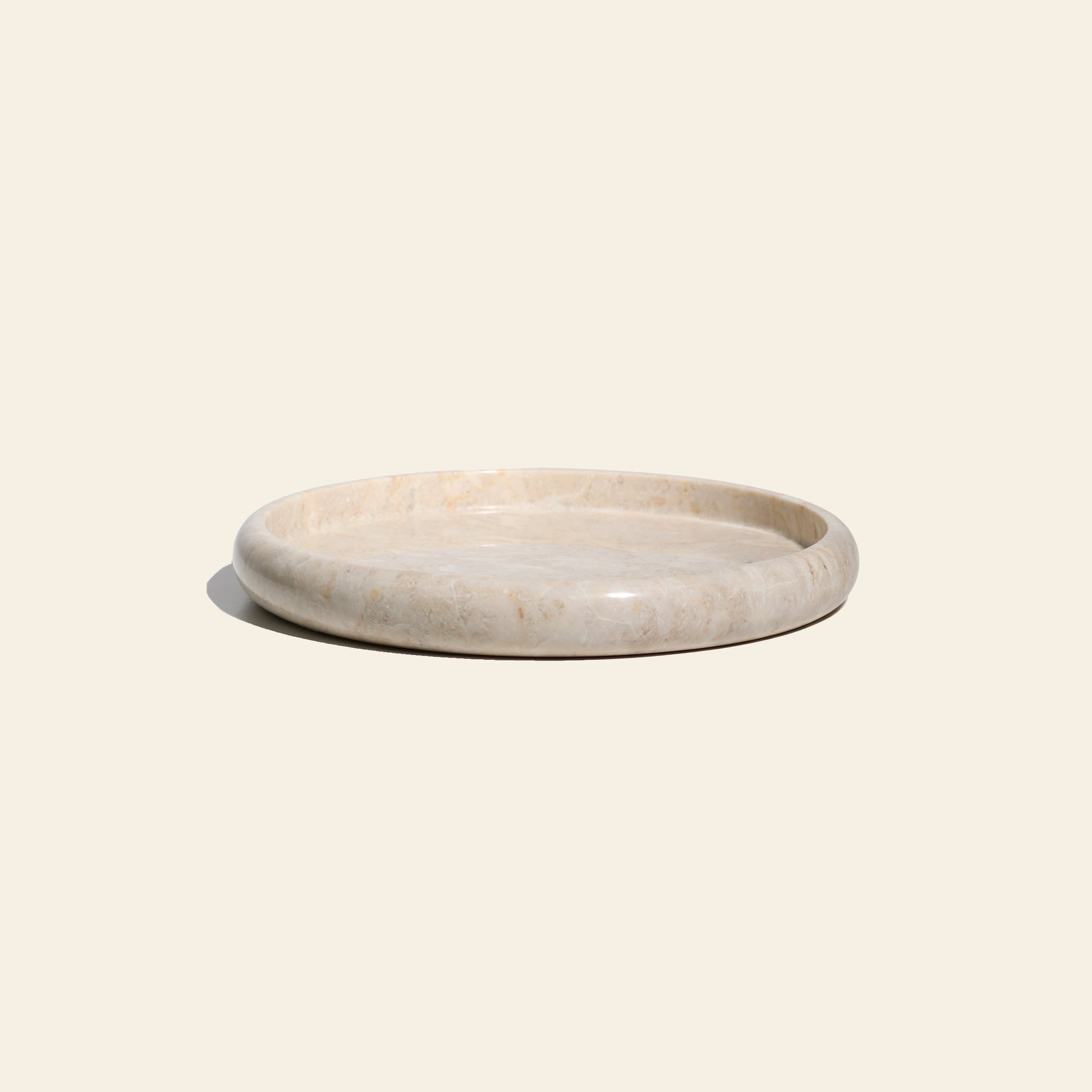The stillgoods Harmony Tray by stillgoods is a round, shallow marble centerpiece with a smooth, polished surface and light beige color, adding subtle elegance to your home decor. Shown on an off-white background.