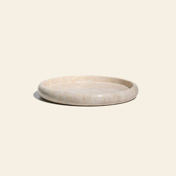 The stillgoods Harmony Tray by stillgoods is a round, shallow marble centerpiece with a smooth, polished surface and light beige color, adding subtle elegance to your home decor. Shown on an off-white background.