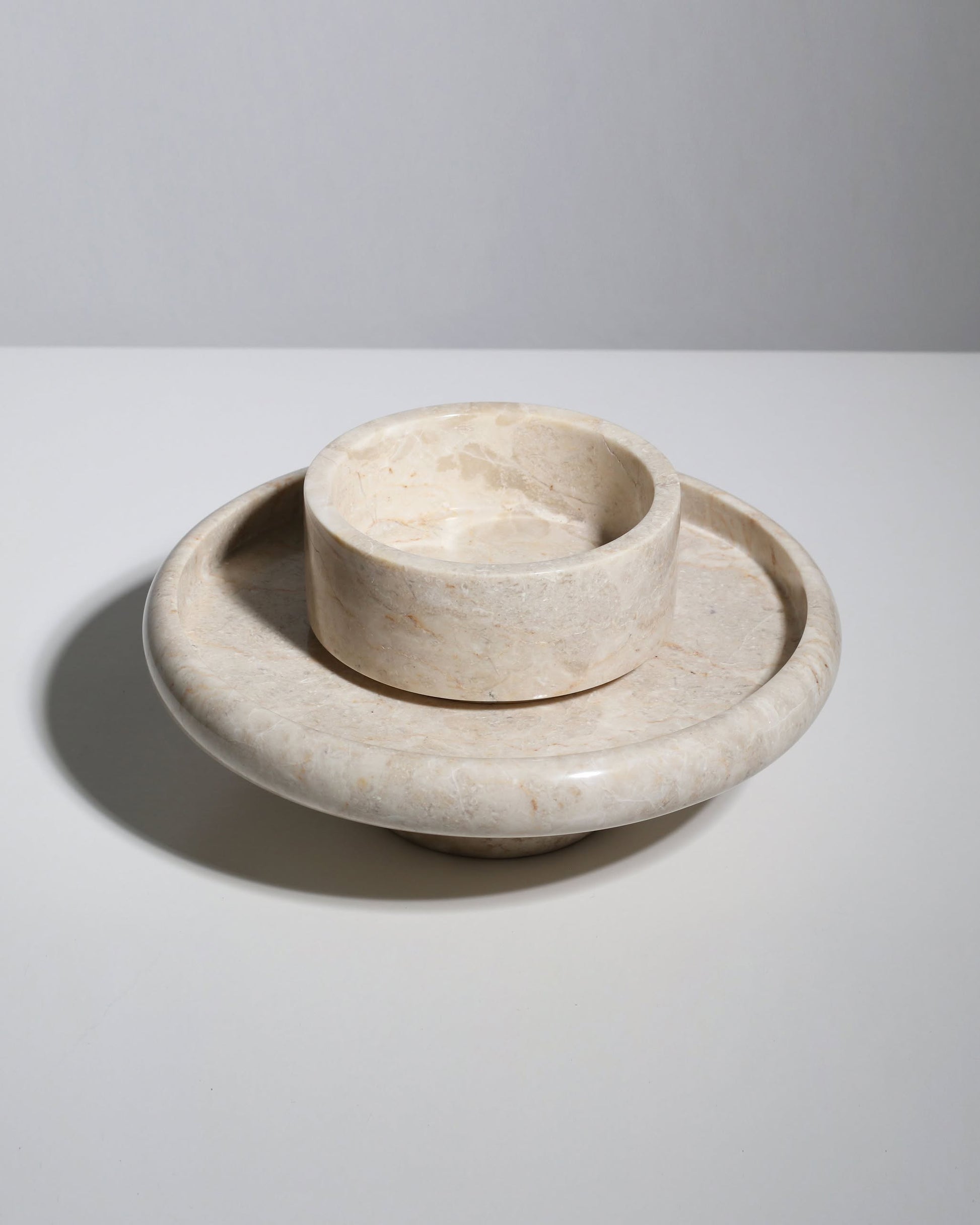 The stillgoods Harmony Tray by stillgoods is a round beige marble centerpiece dish with a raised central bowl and wide flat base, adding elegance to your decor. Shown on a white surface with a plain light gray background.