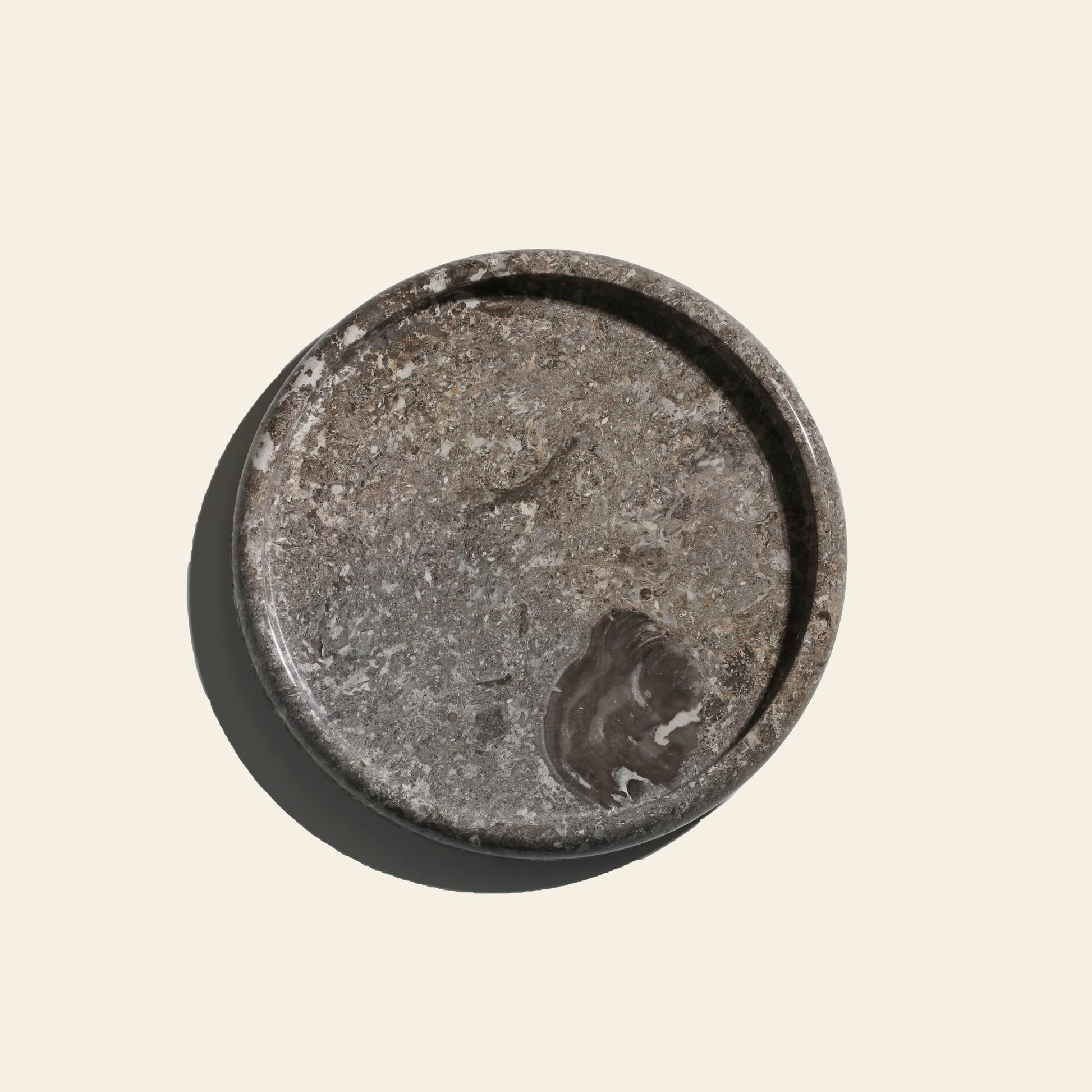 The stillgoods Harmony Tray by stillgoods is a round, flat gray stone tray with marbled texture and white veining, making an elegant centerpiece for home decor. Shown on a light beige background with a shadow on the left.