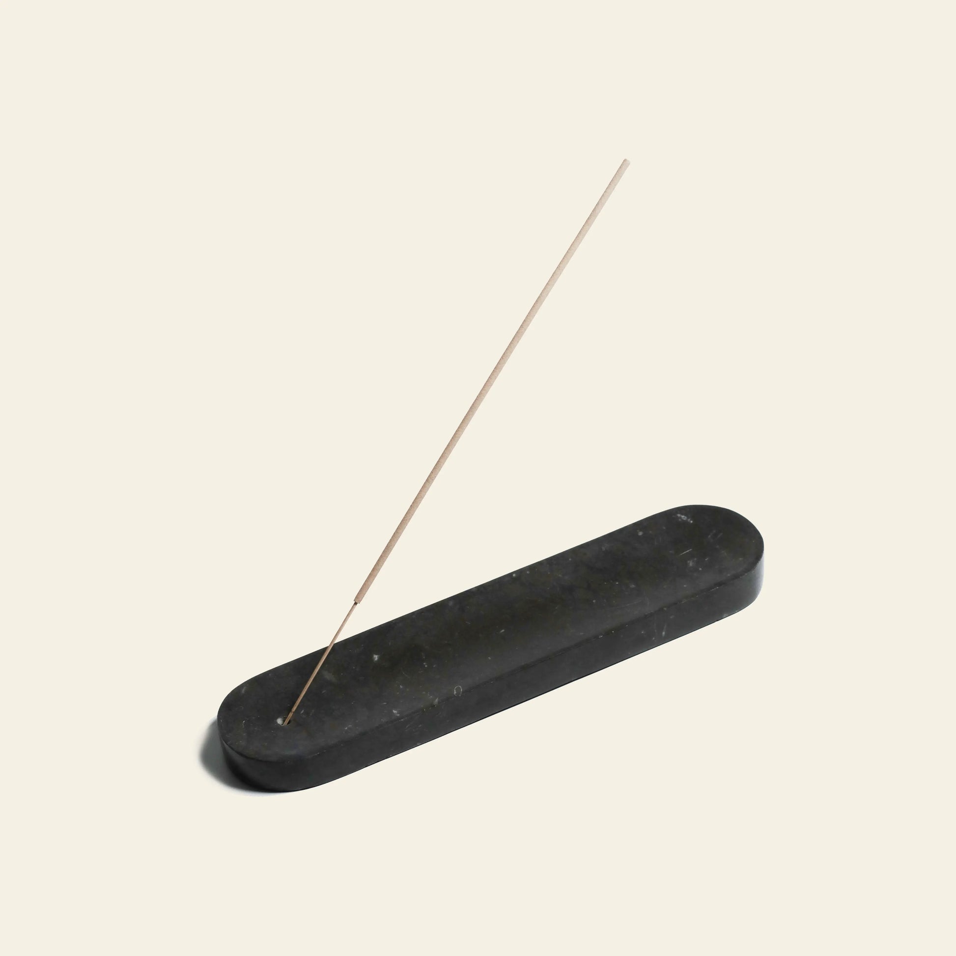 A single lit incense stick with an elongated design rests at an angle in the stillgoods Horizon Incense Holder, a simple black stone piece by stillgoods, displayed on a plain light background.