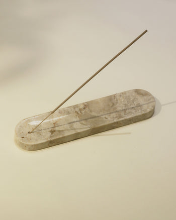 A single lit incense stick rests in the stillgoods Horizon Incense Holder, an elongated marble design by stillgoods, casting a delicate shadow on a light surface.