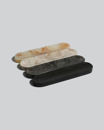 Four smooth, elongated Horizon Incense Holders by stillgoods—cream, light beige, dark gray, and black—are arranged in a row on a plain light gray background. Their versatile design makes them a stylish accent or unique incense holder for any space.