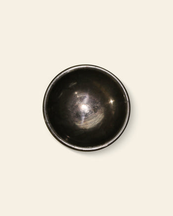 The stillgoods Luna Dish is a small, dark metal bowl with a smooth, rounded shape and a slightly reflective surface, photographed from above against a plain light beige background.