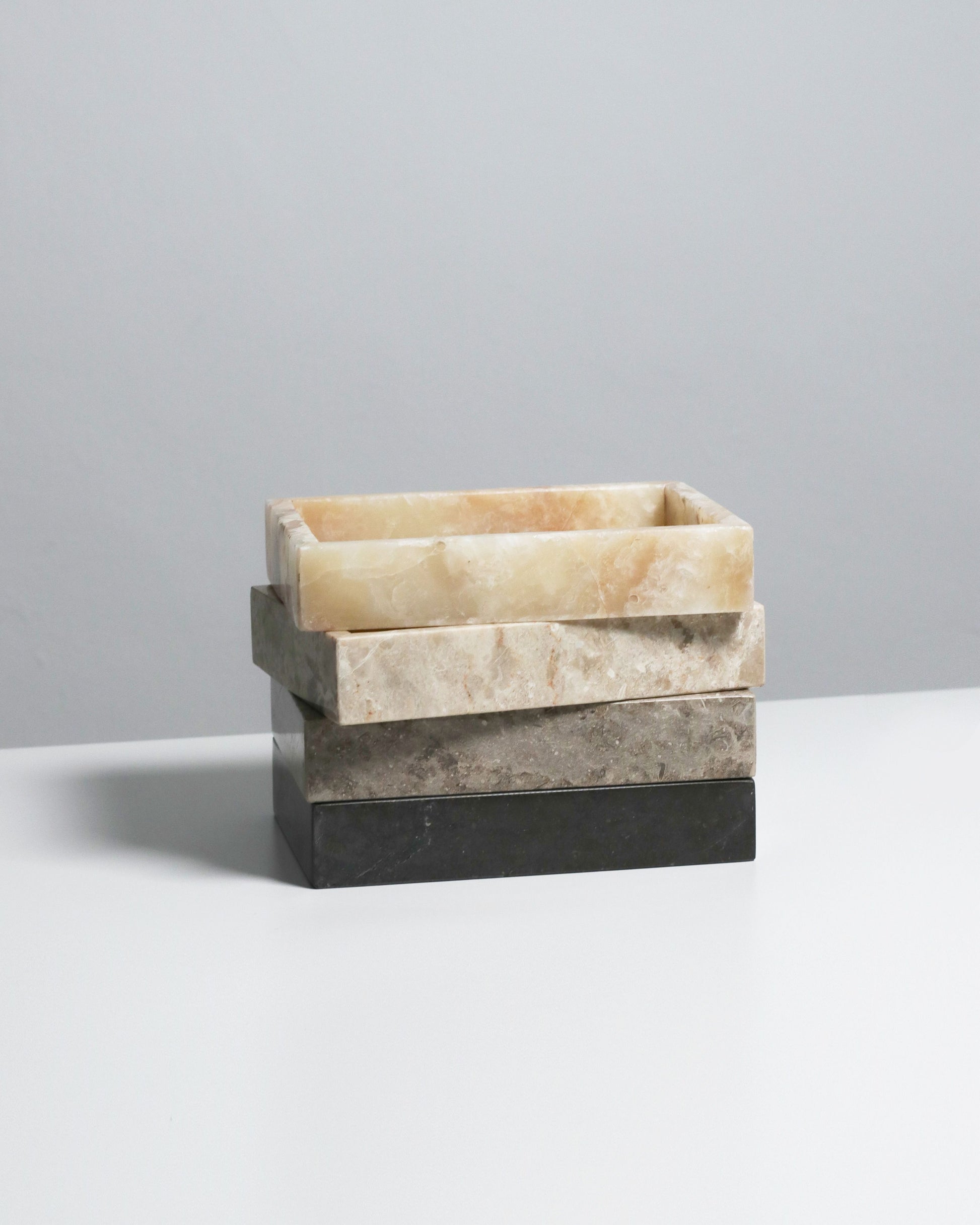 Three stillgoods Mini Valor Trays in cream, beige, and dark gray are stacked slightly askew on a white surface with a light gray background, highlighting their sleek low-profile design for organizing essentials.