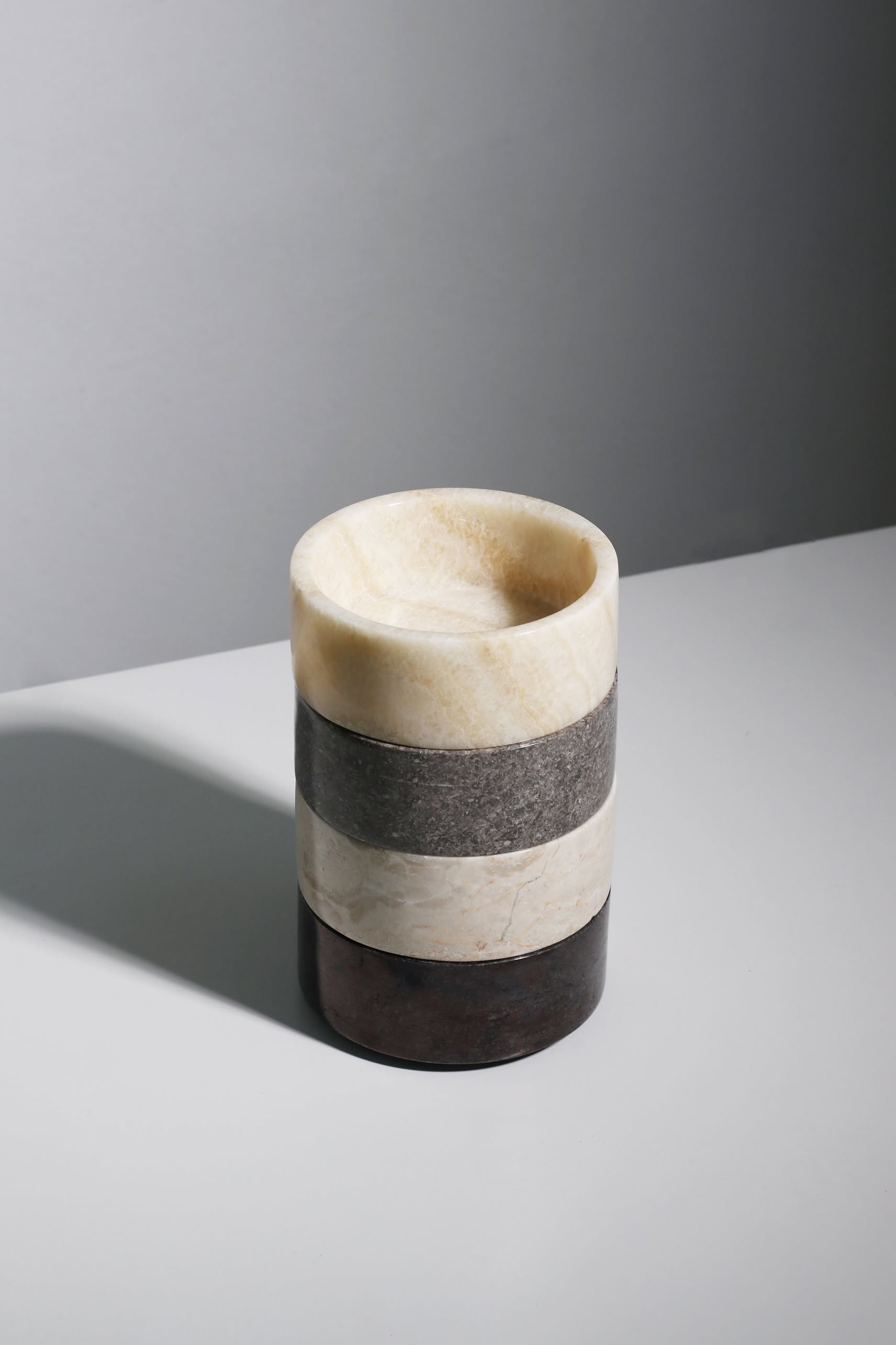 The stillgoods Tingkat Bowl, a cylindrical stone bowl with four horizontal layers in cream, gray, beige, and dark brown onyx-like hues, rests on a light gray surface and casts a gentle shadow to the left.