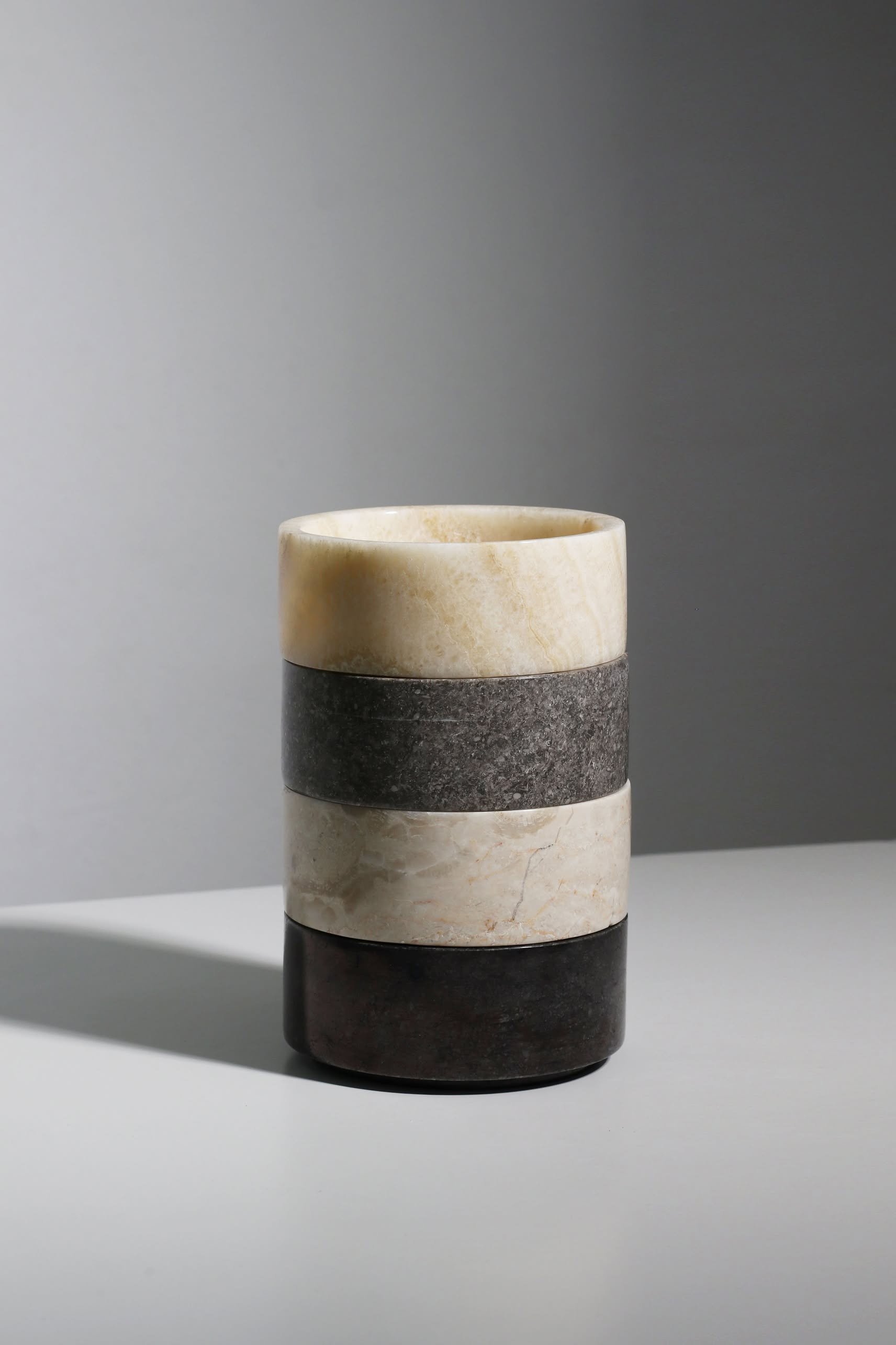 The stillgoods Tingkat Bowl by stillgoods is a cylindrical stone vase with three horizontal bands—light beige, dark gray, and black—resembling marble or onyx, displayed on a light gray surface with a plain gray background.