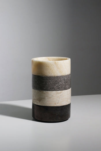 The stillgoods Tingkat Bowl by stillgoods is a cylindrical stone vase with three horizontal bands—light beige, dark gray, and black—resembling marble or onyx, displayed on a light gray surface with a plain gray background.