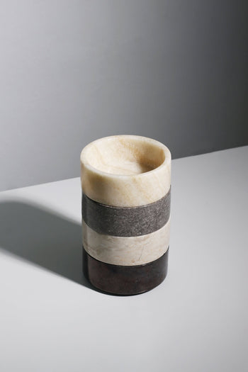 The stillgoods Tingkat Bowl by stillgoods is a cylindrical stone cup with four horizontal layers in beige, gray, and brown marble tones, displayed on a light gray surface against a plain background.