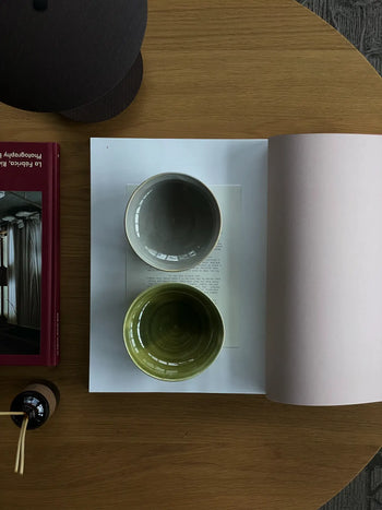 The ves Aki Bowl Set of 2 by ves, featuring gray and green East Asian-inspired ceramic bowls, is displayed on an open book atop a wooden table with a red photography book and stick diffuser partially in view.