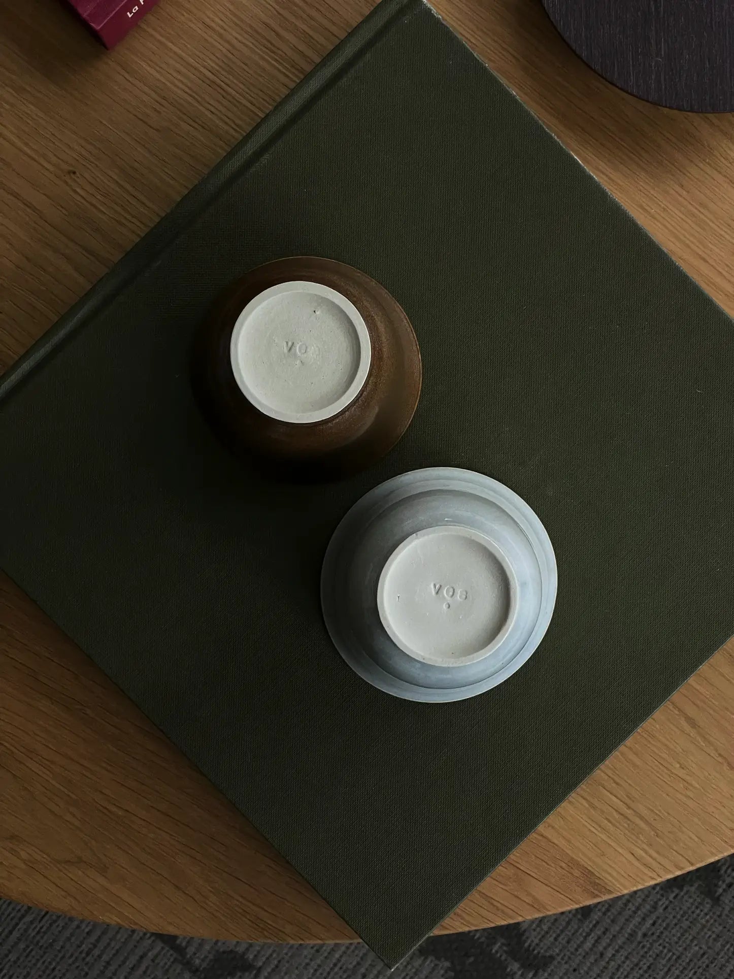 Two ves Aki Bowls by ves—one brown, one light blue—are placed upside down on a dark green book. Inspired by East Asian tableware, both handcrafted ceramic bowls feature YO marked on their bases. Set of 2.