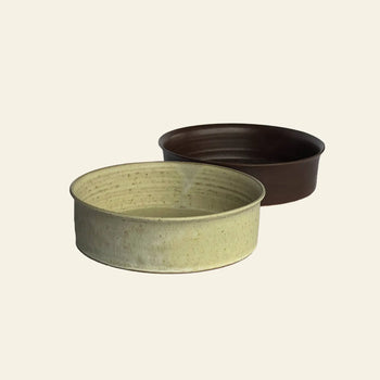 Two round ceramic bowls from the ves Kura Bowl M, Set of 2 are shown on a plain light background. The front bowl is light beige with a speckled finish, while the back one is solid dark brown, highlighting modern handcrafted design.