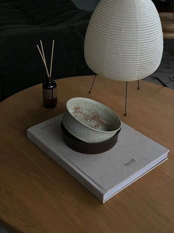 A round wooden table features the ves Kura Bowl M, Set of 2 by ves, handcrafted ceramics, a reed diffuser, and a white paper lantern lamp. A large book titled form lies beneath the bowl, with a dark sofa accenting the modern design.