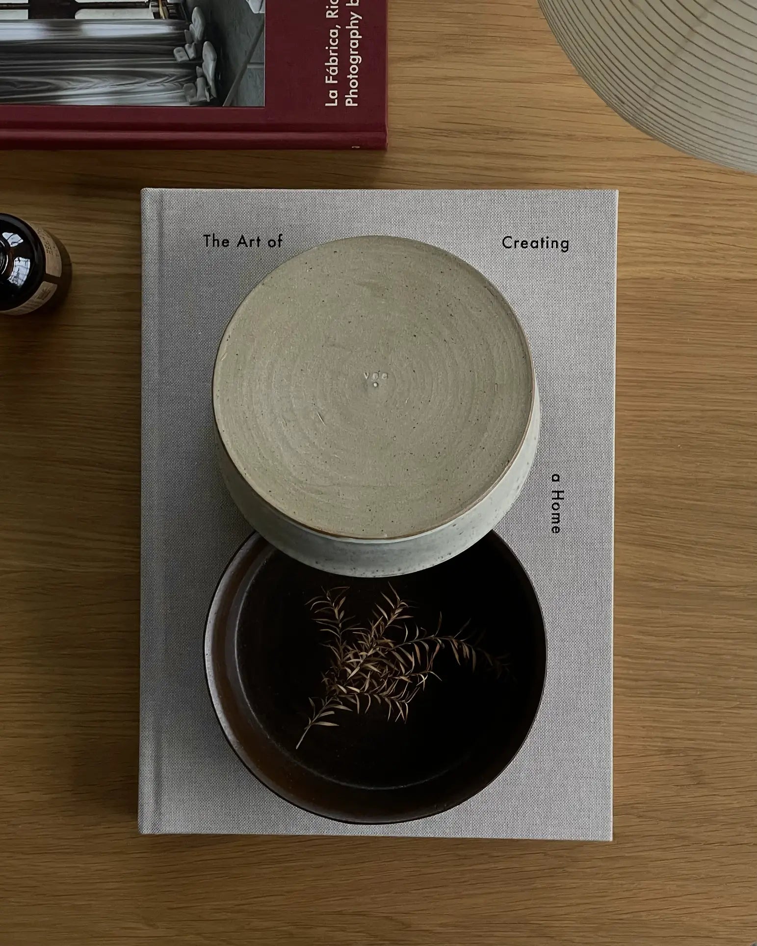 A beige book titled The Art of Creating a Home rests on a wooden table with ves Kura Bowl M, Set of 2 by ves holding dried stems. A lamp, red book, and small brown bottle nearby complete the modern design aesthetic.