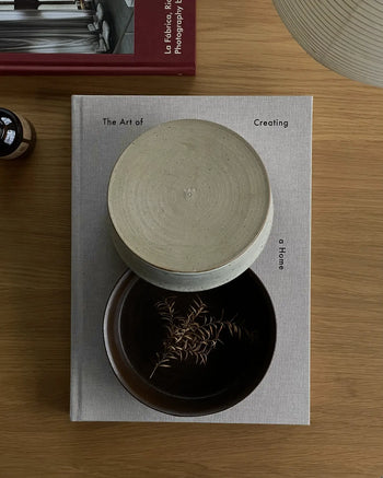 A beige book titled The Art of Creating a Home rests on a wooden table with ves Kura Bowl M, Set of 2 by ves holding dried stems. A lamp, red book, and small brown bottle nearby complete the modern design aesthetic.