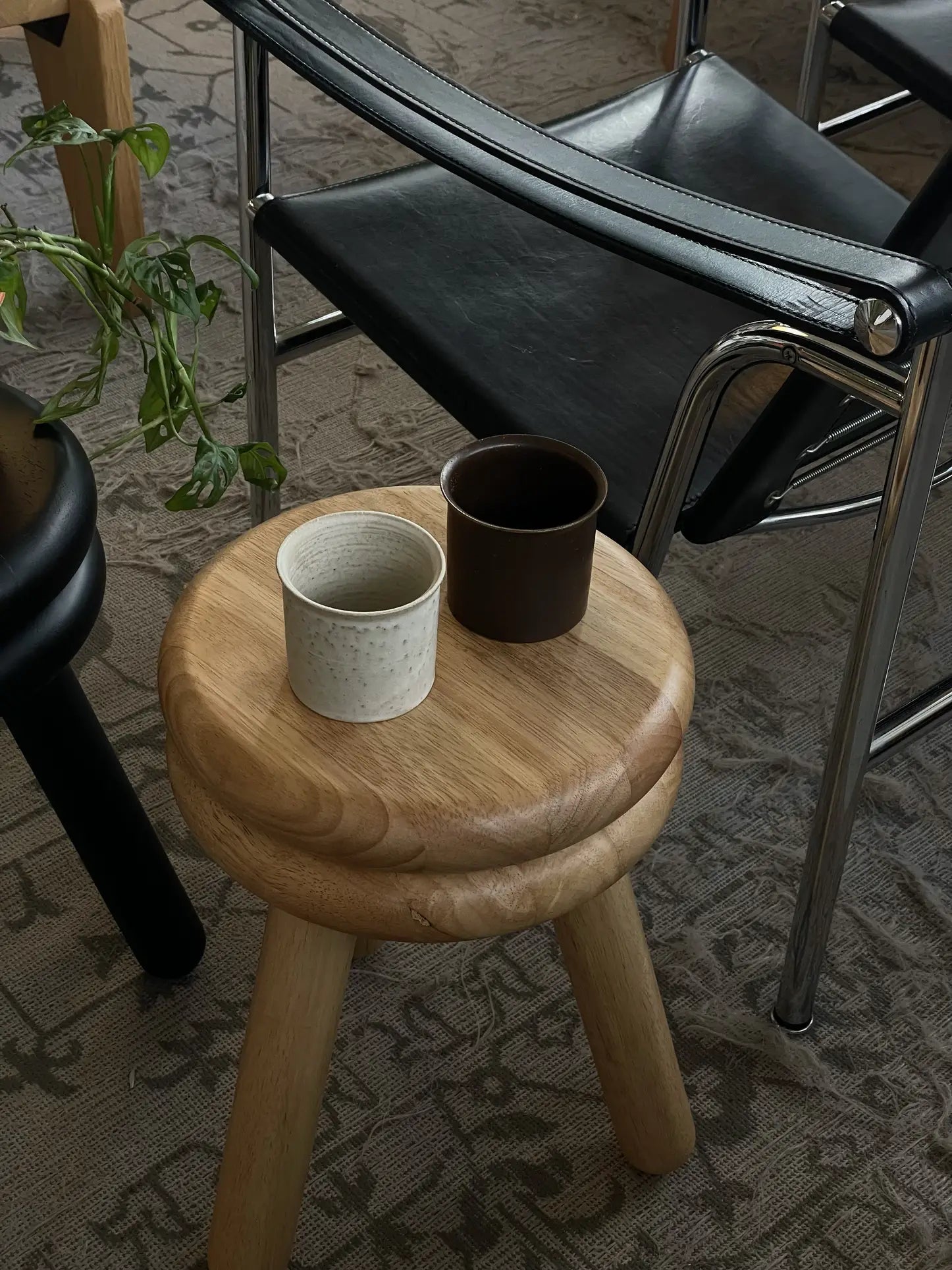 A wooden stool from the ves Kura Collection displays the ves Kura Cup Set of 2—one white, one brown—next to a black metal-framed chair on a patterned carpet, while a green leafy plant brings freshness to this stylish decor scene.